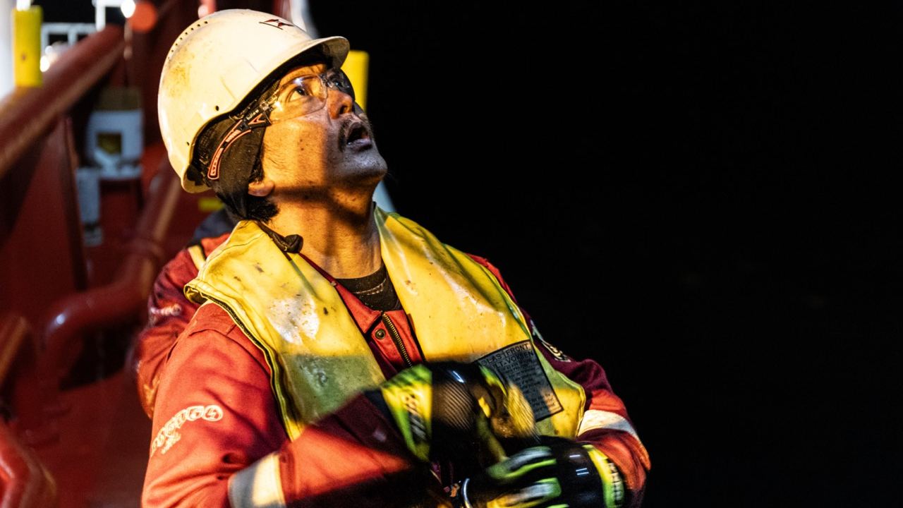 Deck crew member in coveralls, hard hat and life vest watching a lift overhead at night